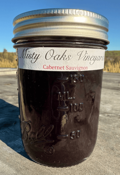 A glass jar filled with cabernet sauvignon jam, labeled Misty Oaks Vineyard Cabernet Sauvignon, outdoors on a wooden surface with a clear blue sky in the background.