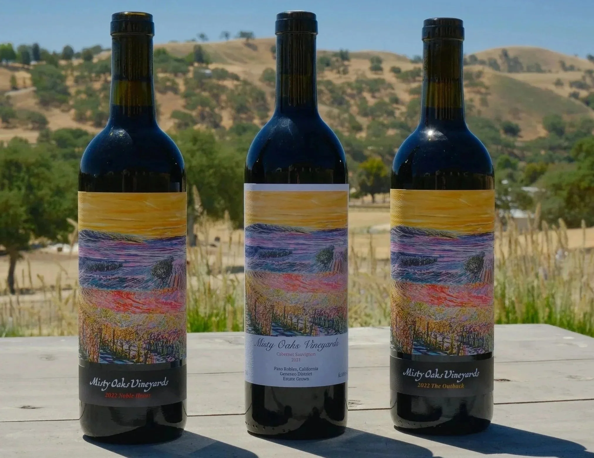 Three bottles of wine from Misty Oaks Vineyards are placed on a wooden surface outdoors. Each bottle has a colorful landscape illustration on its label, with details about the wine types and vintage year. The background features rolling hills, dry grass, and sparse trees under a blue sky.