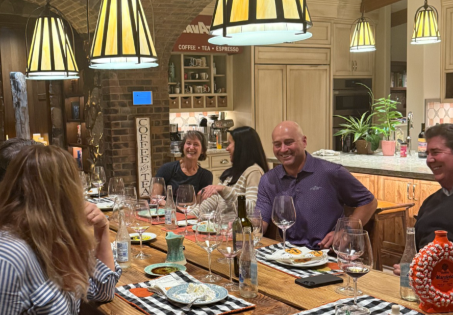 Group of people sitting at a dining table in a cozy, warmly lit kitchen or dining area, laughing and enjoying a meal together with wine glasses, bottles, and plates on the table.