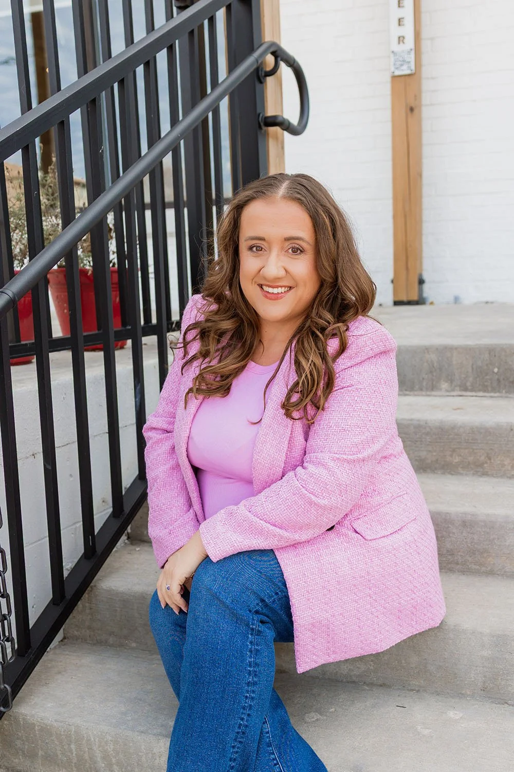 A woman with long, wavy brown hair sitting on outdoor concrete stairs, wearing a pink blazer and blue jeans, smiling at the camera.