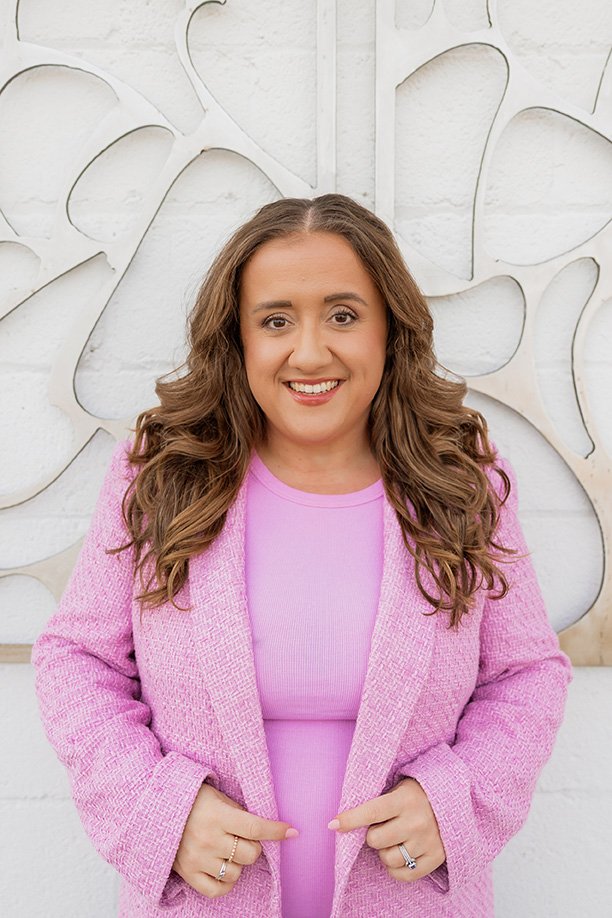 A woman with brown wavy hair, wearing a pink blazer and a pink top, smiling and standing in front of abstract wall art.