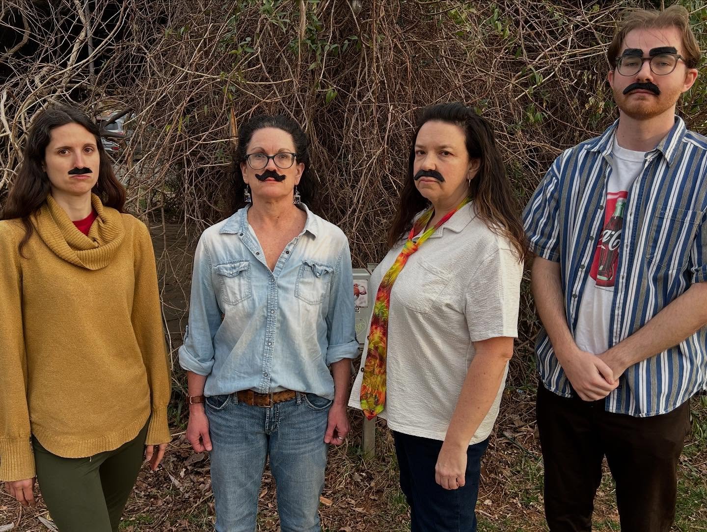 Four people standing outdoors in front of a dense tangle of dry vine-covered branches, each wearing a fake mustache, with serious expressions. Improv class Charlottesville comedy Decipher Potter's fun date night Bent Theatre