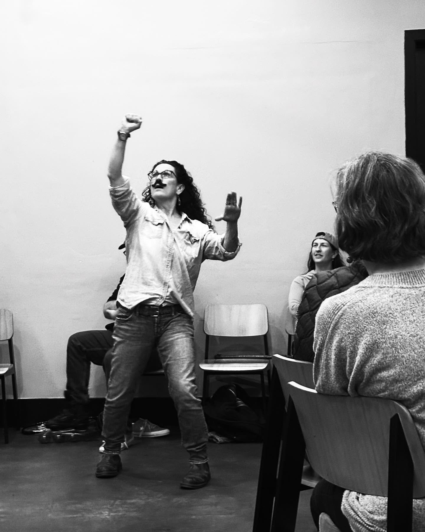 A woman with curly hair and glasses is standing and gesturing with her right arm raised and her left arm slightly out. Improv class Charlottesville comedy Decipher Potter's fun date night Bent Theatre
