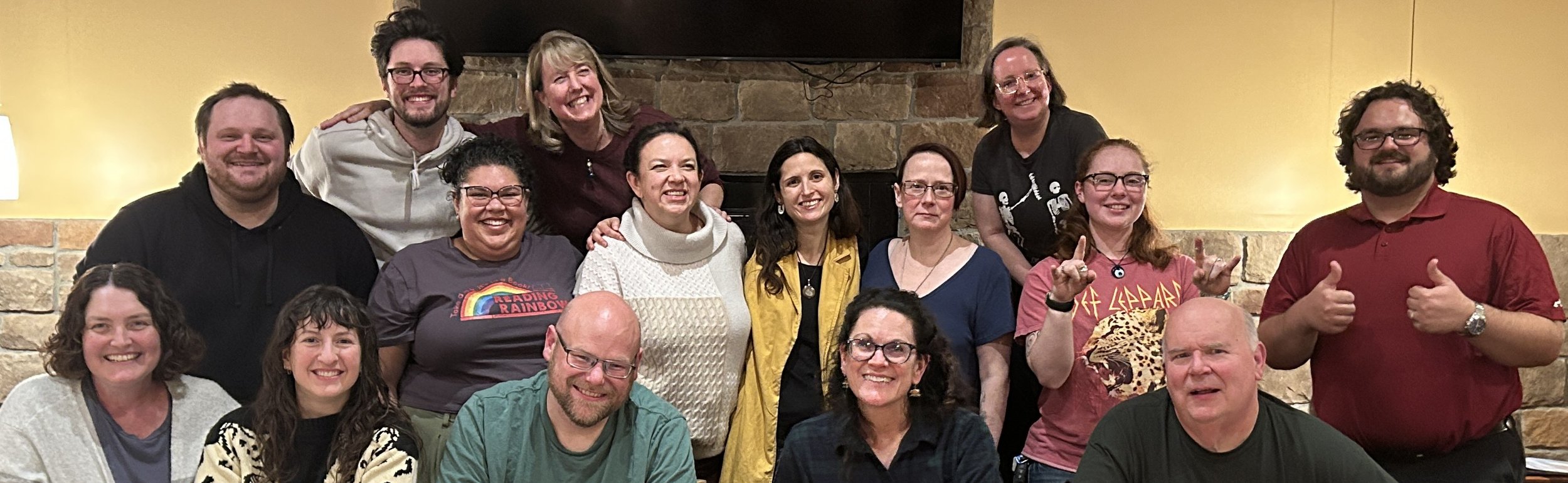 Group of 16 people posing together indoors in front of a stone fireplace, smiling and making various gestures. Improv class Charlottesville comedy Decipher Potter's fun date night Bent Theatre