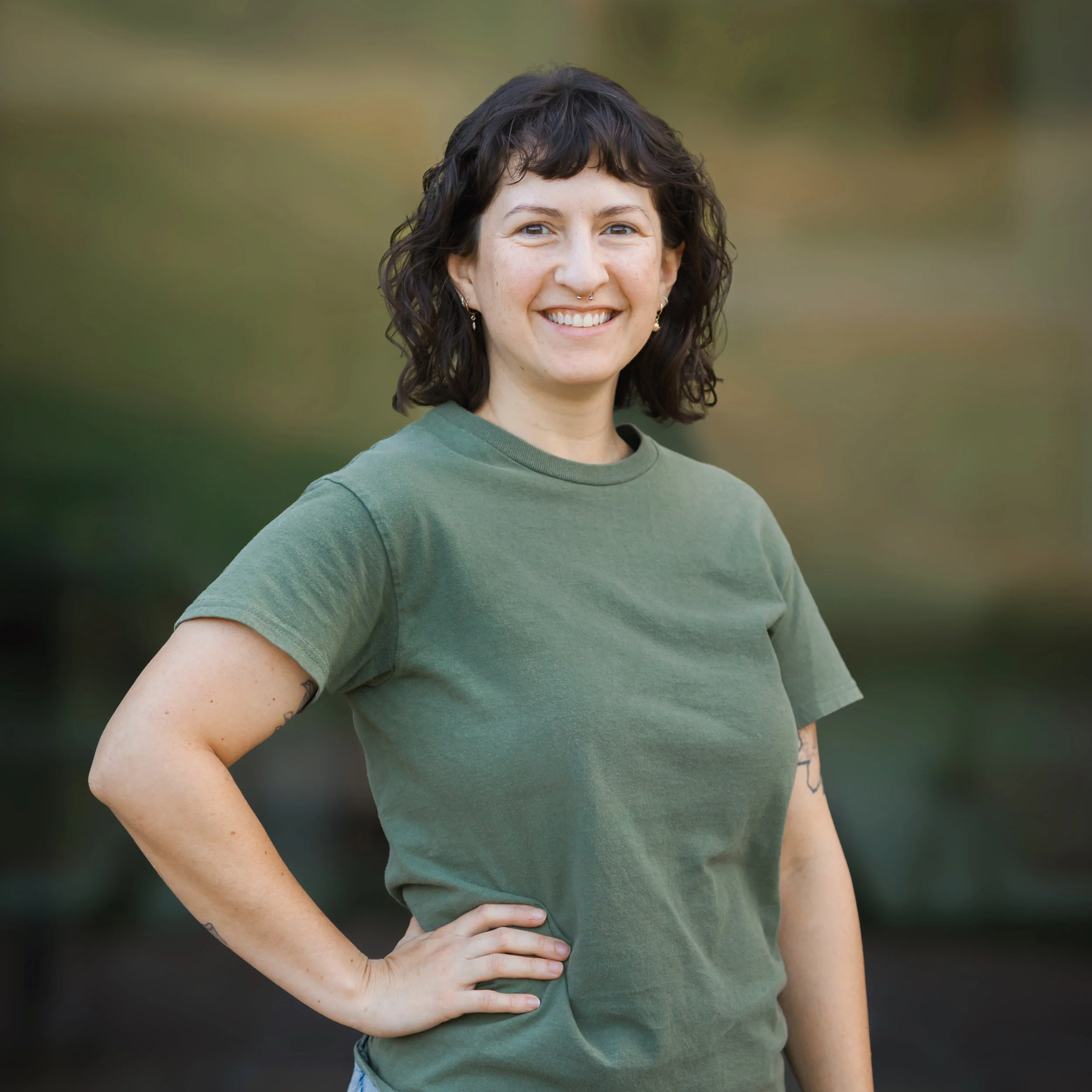 Sophie Elliott with shoulder-length dark hair standing against a plain brown wall, wearing a beige long sleeve shirt and black overalls. Improv Charlottesville comedy Decipher Potter's fun date night