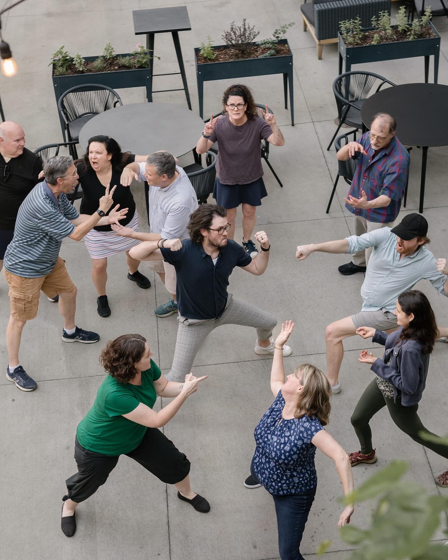 A group of diverse people engaging in a lively, playful fight or dance on an outdoor patio with tables and potted plants.