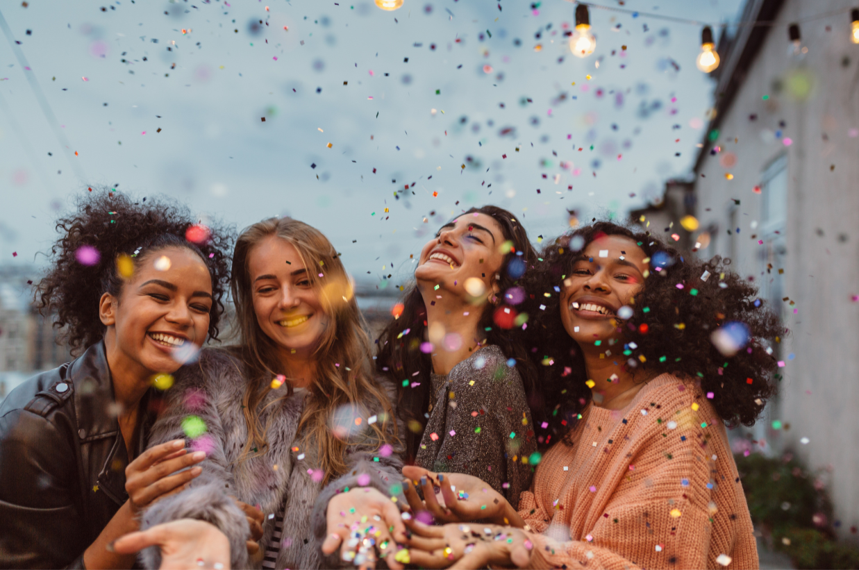 Four women celebrating with colorful confetti falling around them on an outdoor rooftop. They are smiling and enjoying the moment.