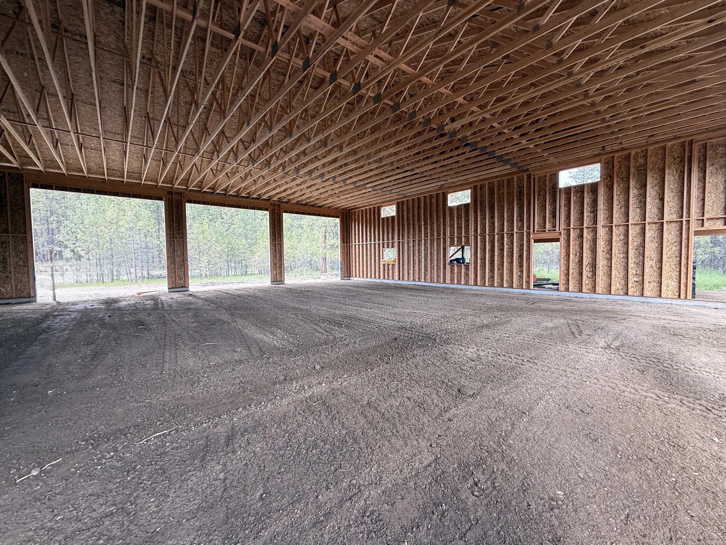 The interior of a building under construction with exposed wooden framing, open walls, and a gravel floor, overlooking a green forested area outside. Sub grade for concrete foundation.