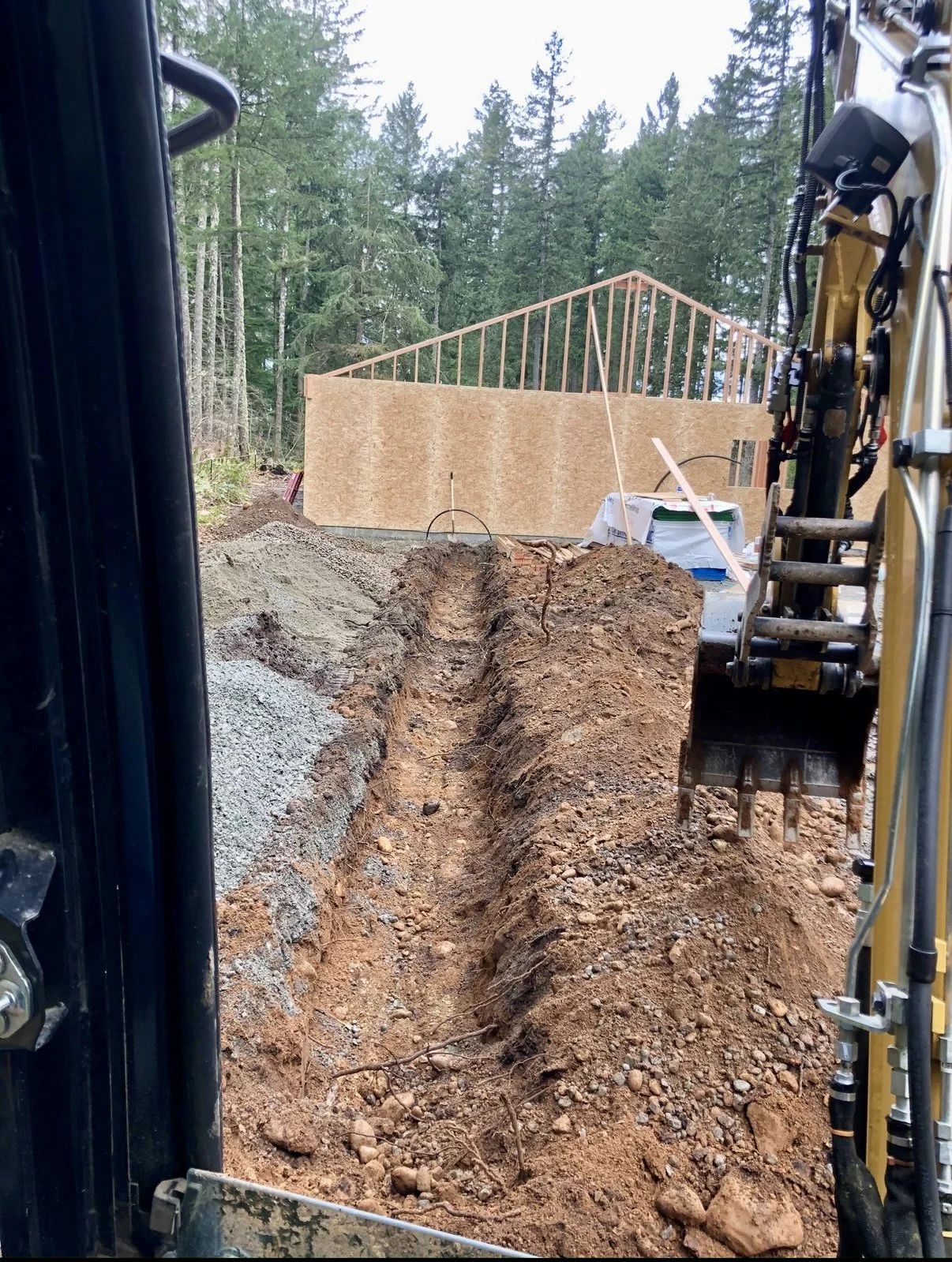 Construction site with dug trench, wooden framing for building wall, and forest in background.