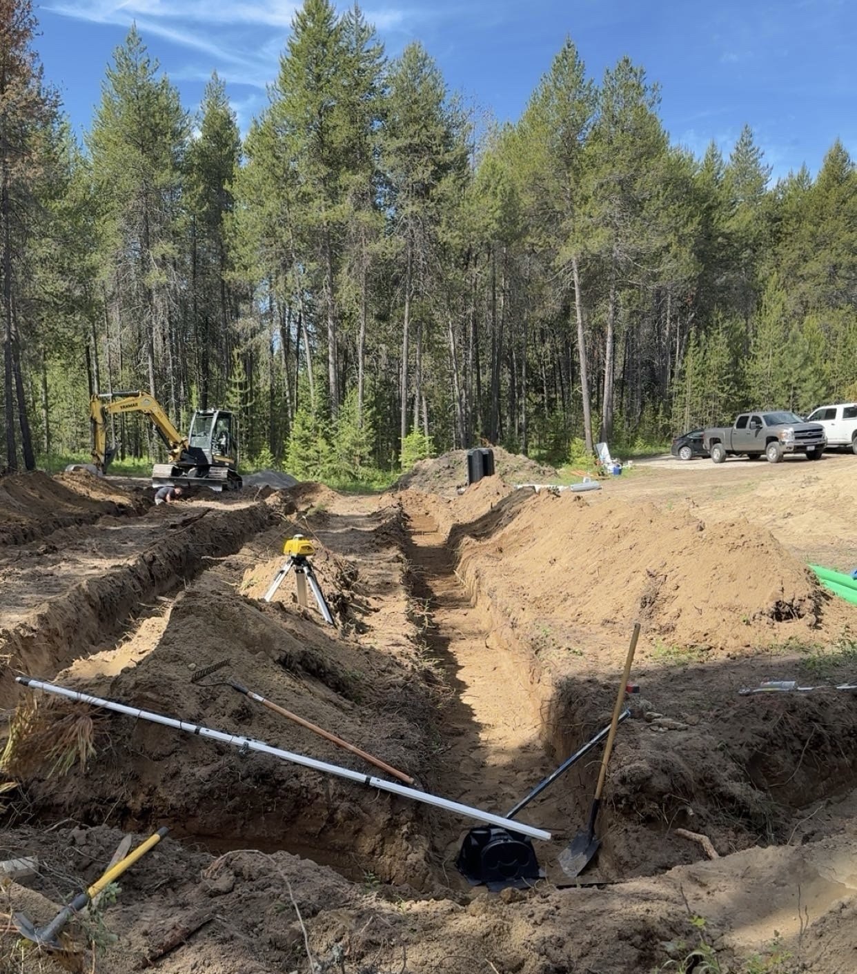 Construction site with a trench being dug, a small excavator, surveying equipment, and parked trucks in a wooded area on a sunny day.