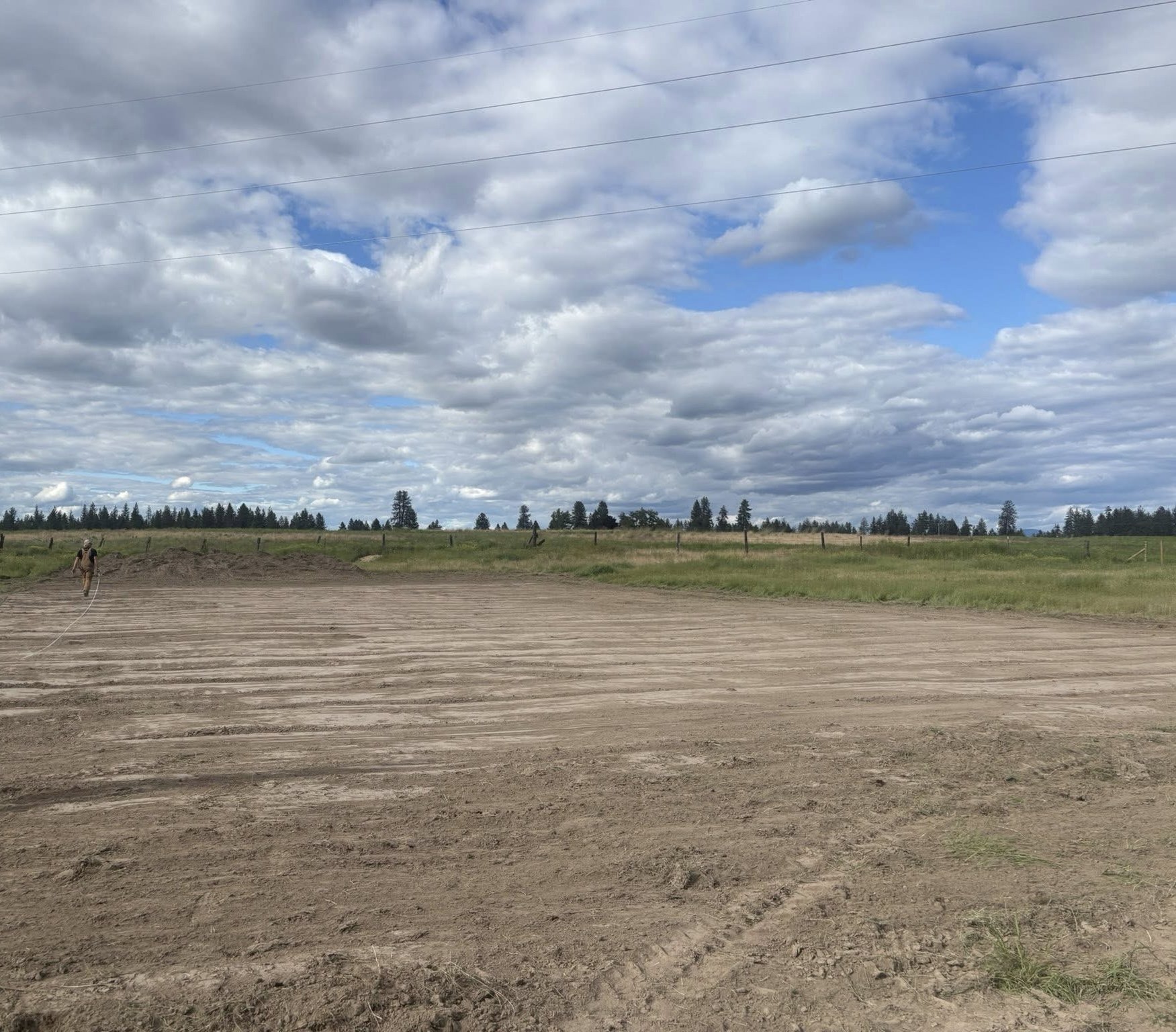 An open dirt field with parallel lines marking the soil, surrounded by grassy land with trees in the background and a partly cloudy sky overhead.