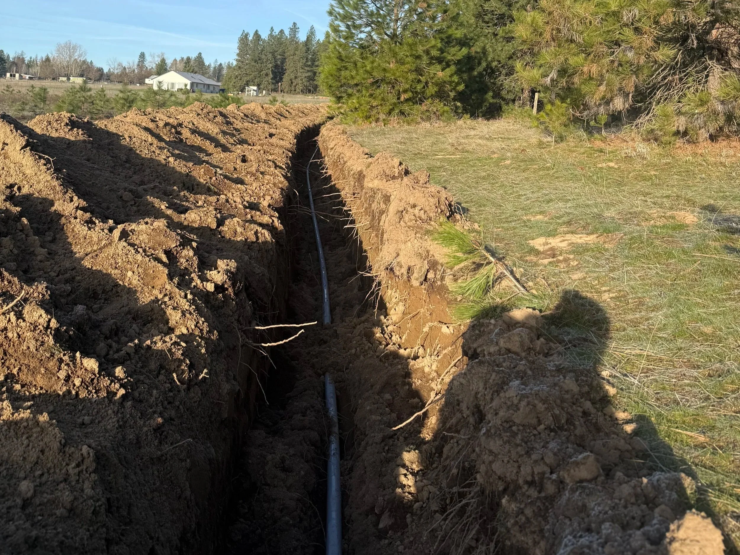 A construction site with a trench dug in the ground and a black conduit pipe running along the bottom. There are trees and houses in the background, and the sky is clear.