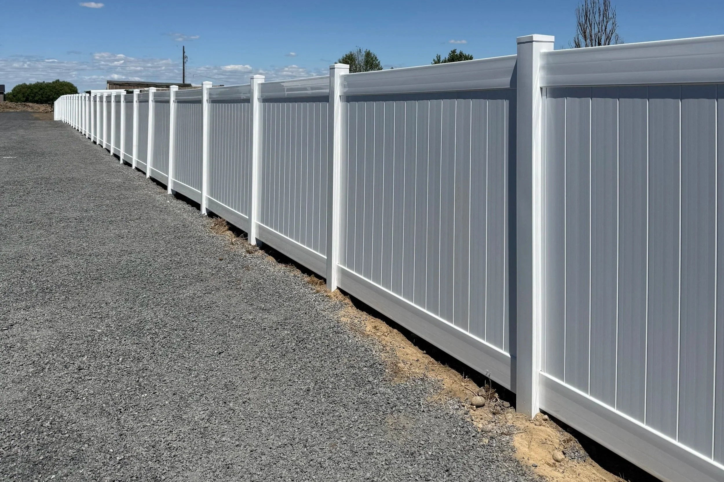 A long white vinyl privacy fence running along a gravel surface on a sunny day with a partly cloudy sky.