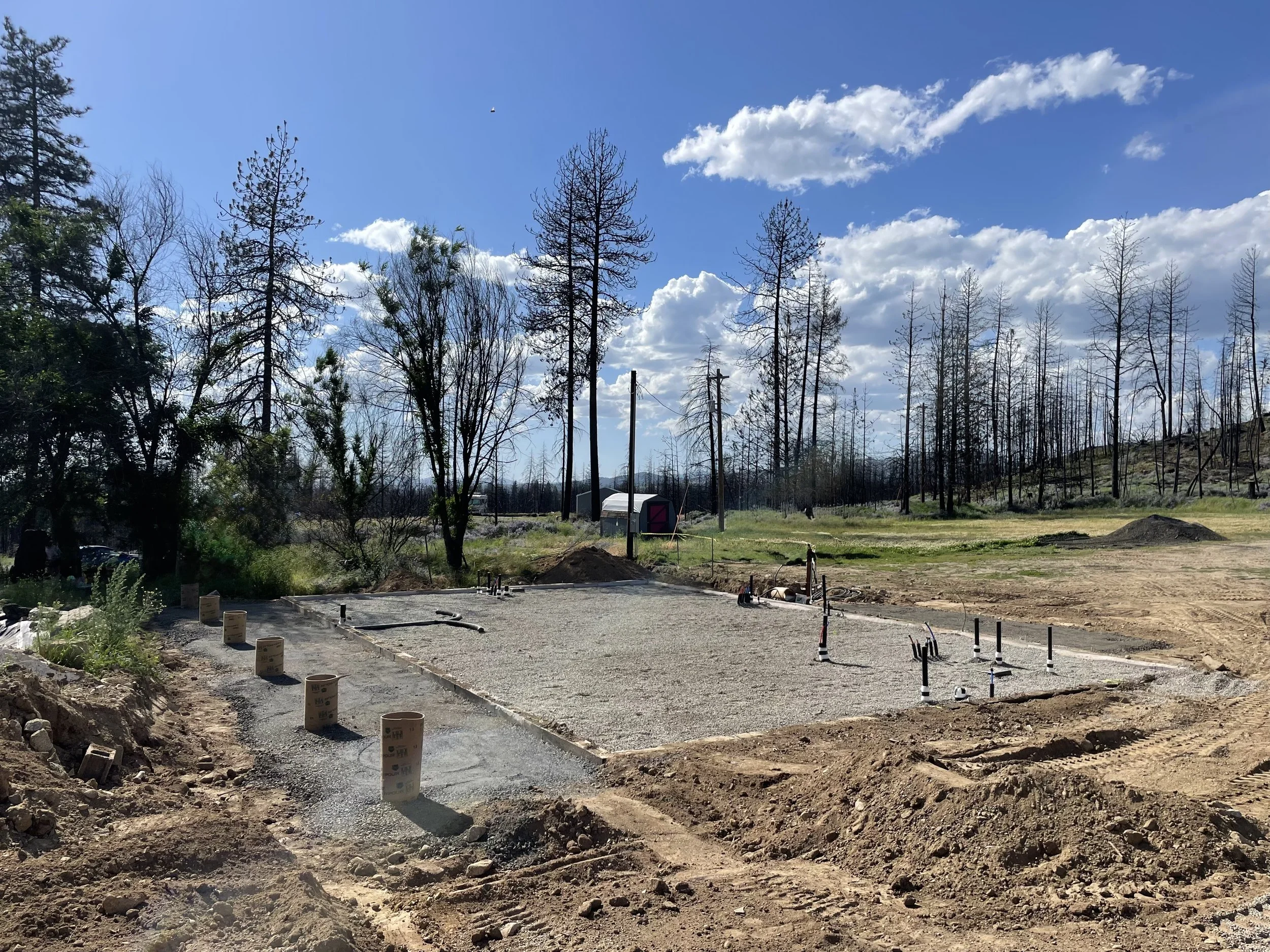 A construction site with freshly poured concrete, surrounded by dirt and construction materials, and trees with a blue sky and scattered clouds in the background.