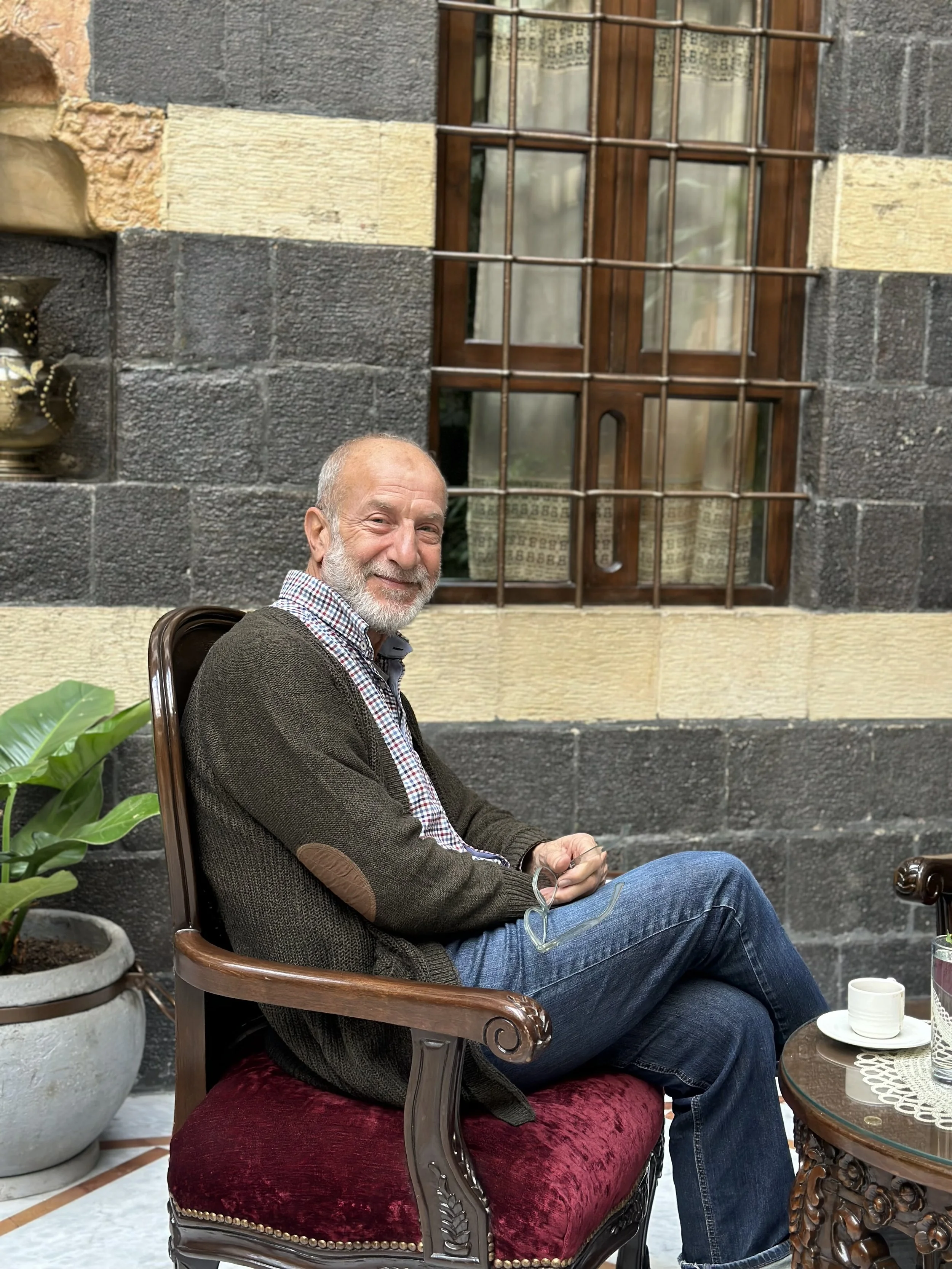 An older man with a beard sitting on a vintage-style wooden chair with a red velvet cushion, smiling, next to a potted green plant, with a building featuring stone and brick walls and a window with wooden bars in the background.