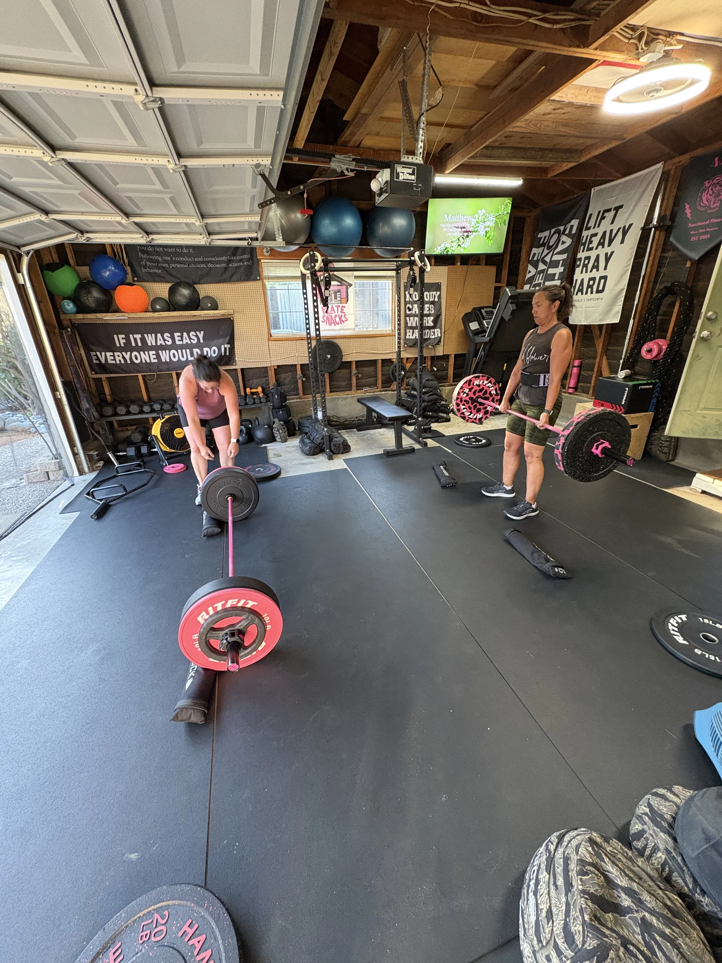 Two women exercising in a home gym. One is lifting a kettlebell, and the other is doing a plank on a yoga mat. Various workout equipment and a whiteboard with exercises are visible in the background.
