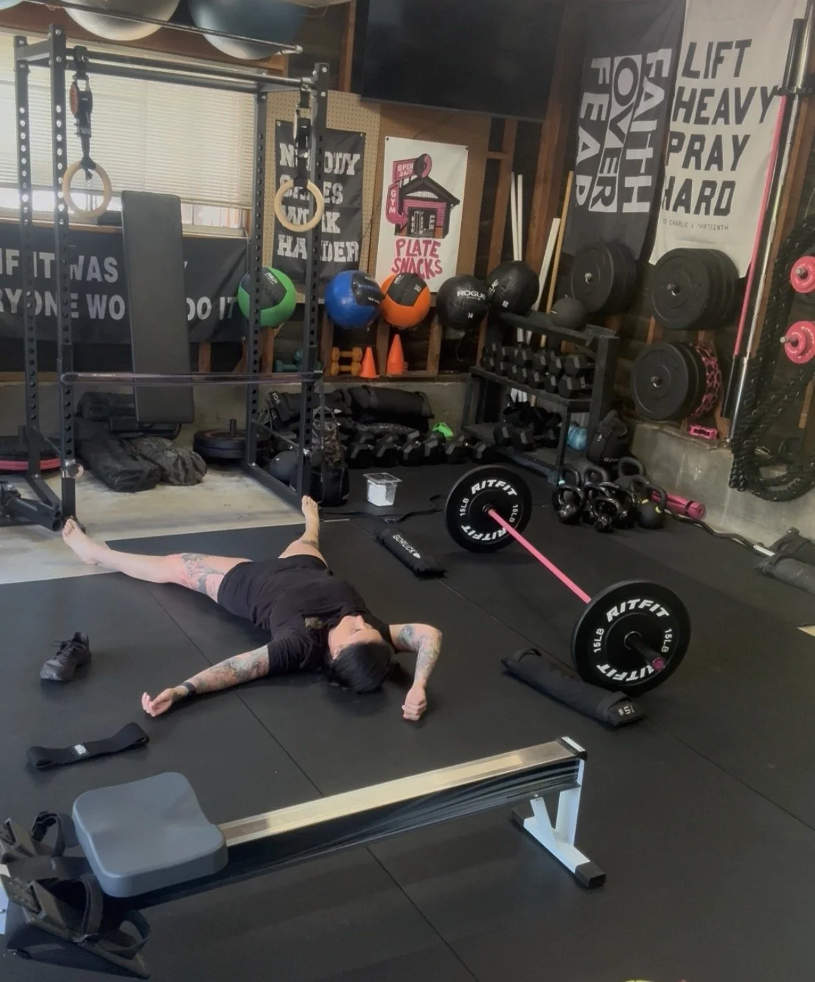 Home gym setup in a garage with fitness equipment, including a barbell with weights, battle ropes, exercise mats, and a stability ball. An open garage door reveals an outdoor view with greenery.