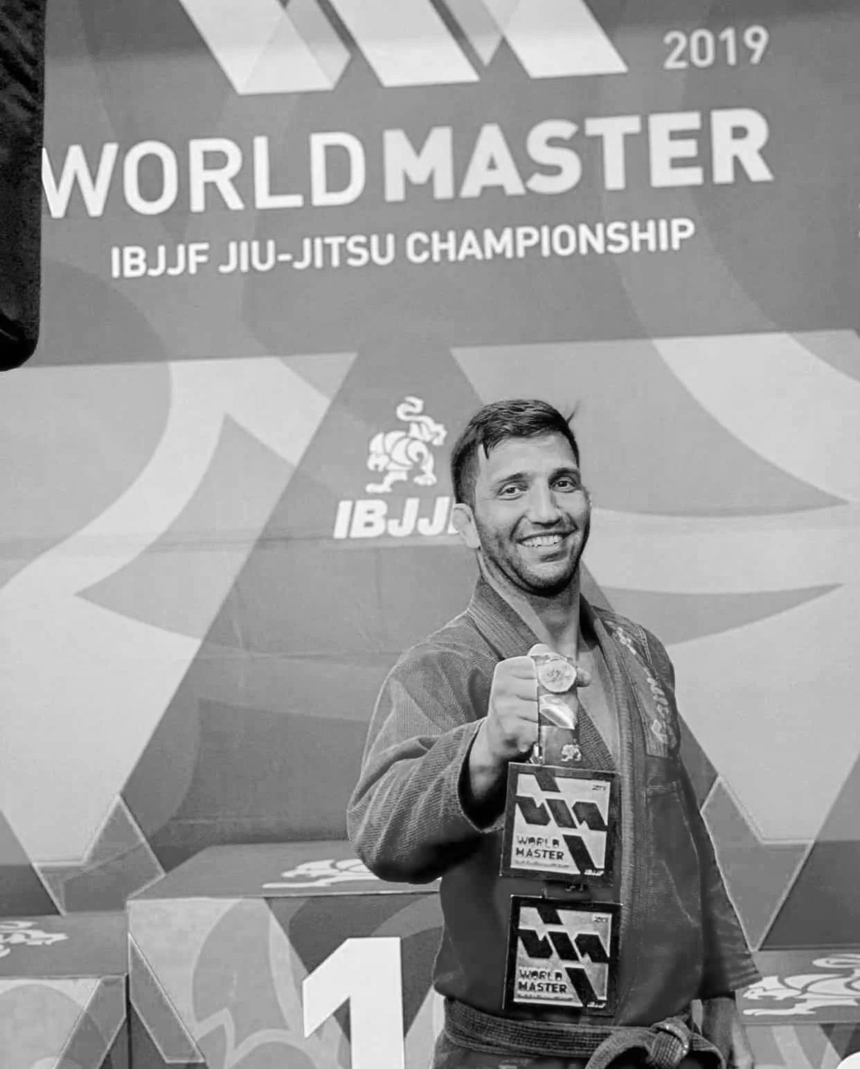 A man in a Brazilian Jiu-Jitsu gi holding medals in front of a backdrop for the 2019 IBJJF World Master Jiu-Jitsu Championship.