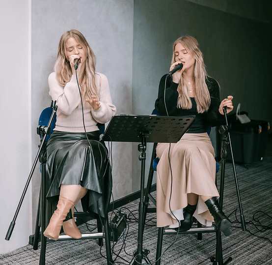 Two women sitting and singing into microphones with closed eyes, wearing long skirts and sweaters in a studio setting.