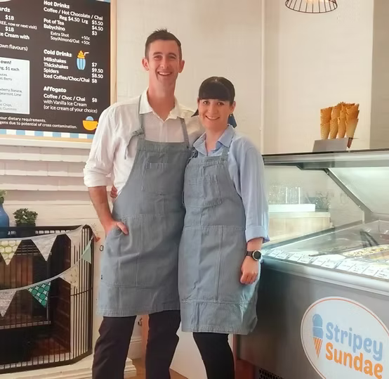 Two people wearing aprons stand inside an ice cream shop, with a display freezer and a menu board visible in the background.