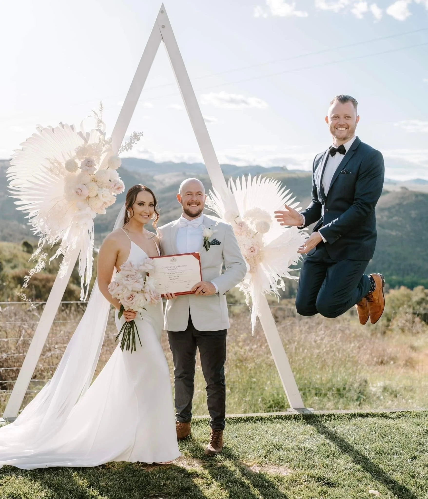 Bride and groom holding a certificate, standing outdoors in front of a triangle arch with floral decorations; a person in a suit is jumping beside them.