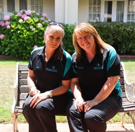 Two women sitting on a bench outdoors with a garden background, both wearing matching black and teal uniforms, smiling at the camera.