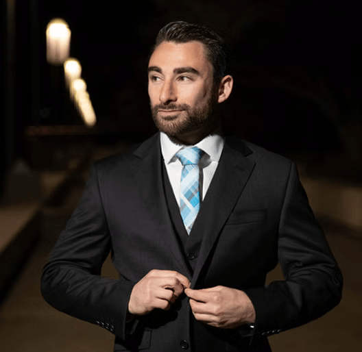 Man in a suit with a blue patterned tie standing outside at night, adjusting his jacket, with streetlights in the background.