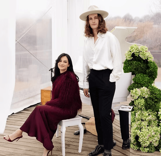 A man in a white shirt and hat stands beside a woman seated in a red outfit, with a floral display shaped like an 'S' in the background.