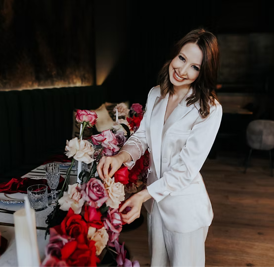 A woman in a white suit setting a floral arrangement on a dining table.