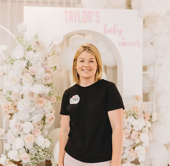 Woman in front of floral decorations at a baby shower with a sign reading "Taylor's Baby Shower."