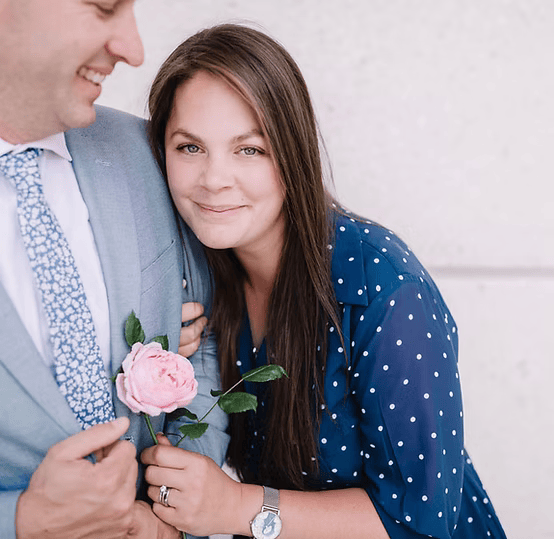 A woman in a blue polka dot dress smiling and holding a pink rose, standing next to a man in a light gray suit and blue tie.