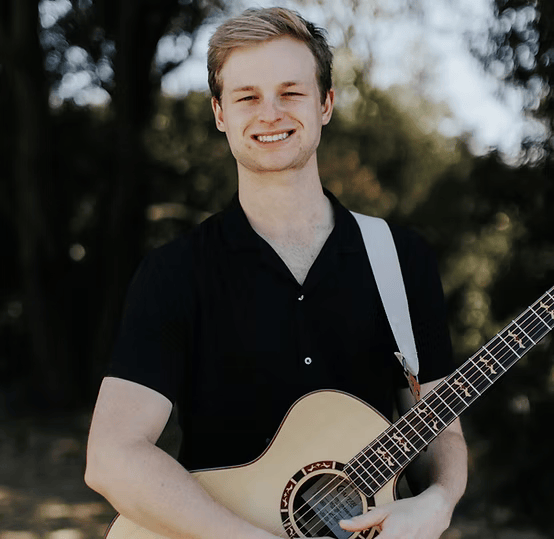Person holding a guitar outdoors with trees in the background, smiling.