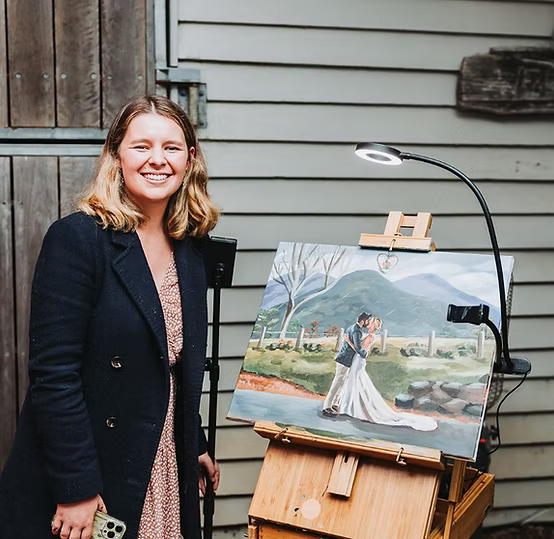 Woman smiling next to an easel displaying a wedding painting, under overhead light.