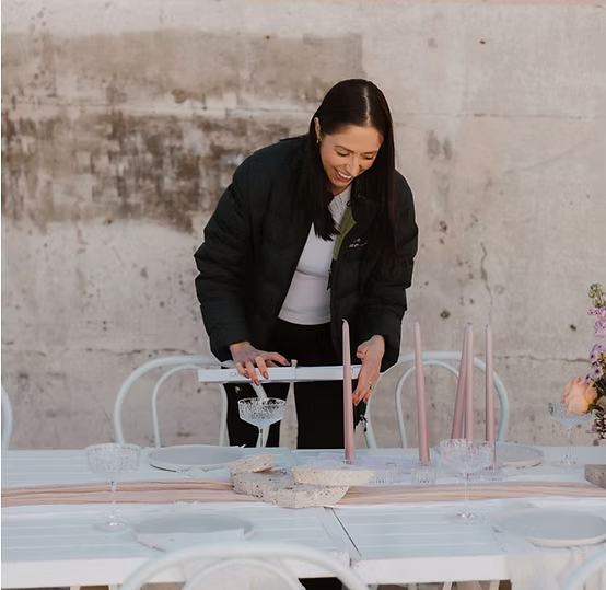 A woman setting up a table outdoors, arranging pink candles and crystal glassware on a white table with decorative flowers.