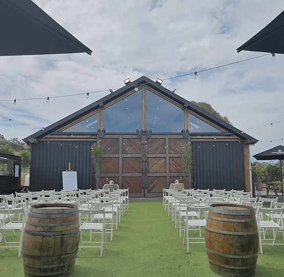 Outdoor wedding venue with a rustic barn backdrop, white chairs, string lights, and wine barrels on a grassy aisle.