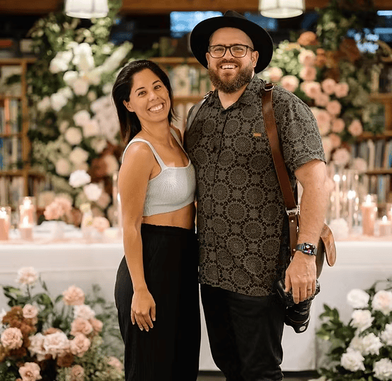 Two people smiling in front of a floral backdrop, with candles and flowers in the background of a decorated setting, possibly for an event or celebration.