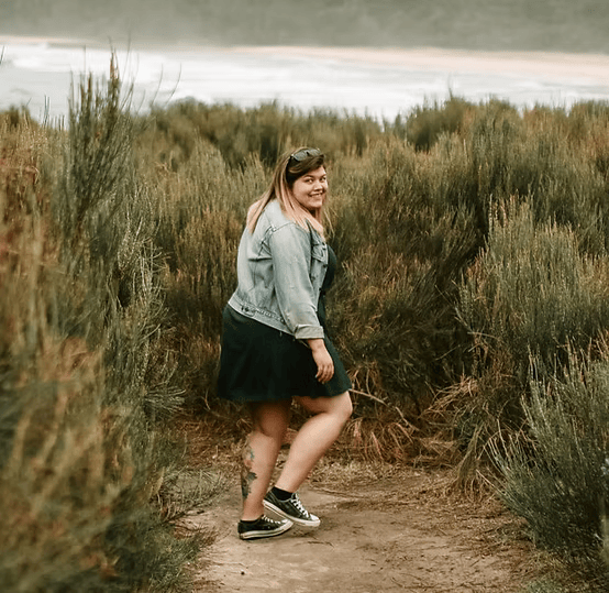 A person walking on a sandy path surrounded by tall grasses, wearing a denim jacket, black dress, and sneakers. The background shows a blurred view of water and distant landscape.