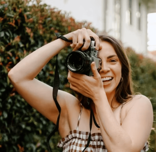 Woman smiling and holding a camera, taking a photo outdoors with greenery in the background.