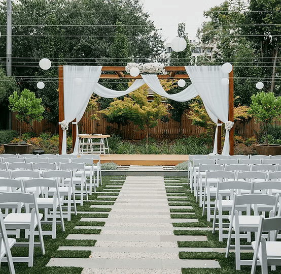 Outdoor wedding ceremony setup with white chairs and arbor draped in white fabric and flowers