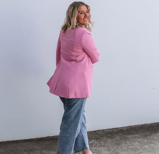 Person wearing a pink blazer and blue jeans standing and smiling