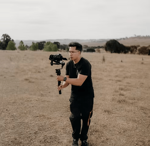 Person holding a camera stabilizer in a dry grassy field, with trees in the background.
