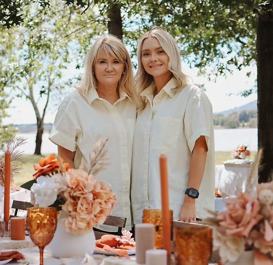 Two women standing outdoors near a decorated table with flowers and candles, wearing matching light-colored shirts.