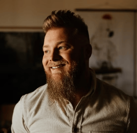Smiling man with a beard and short hair, wearing a light-colored shirt, looking to the side, in a warmly lit room.