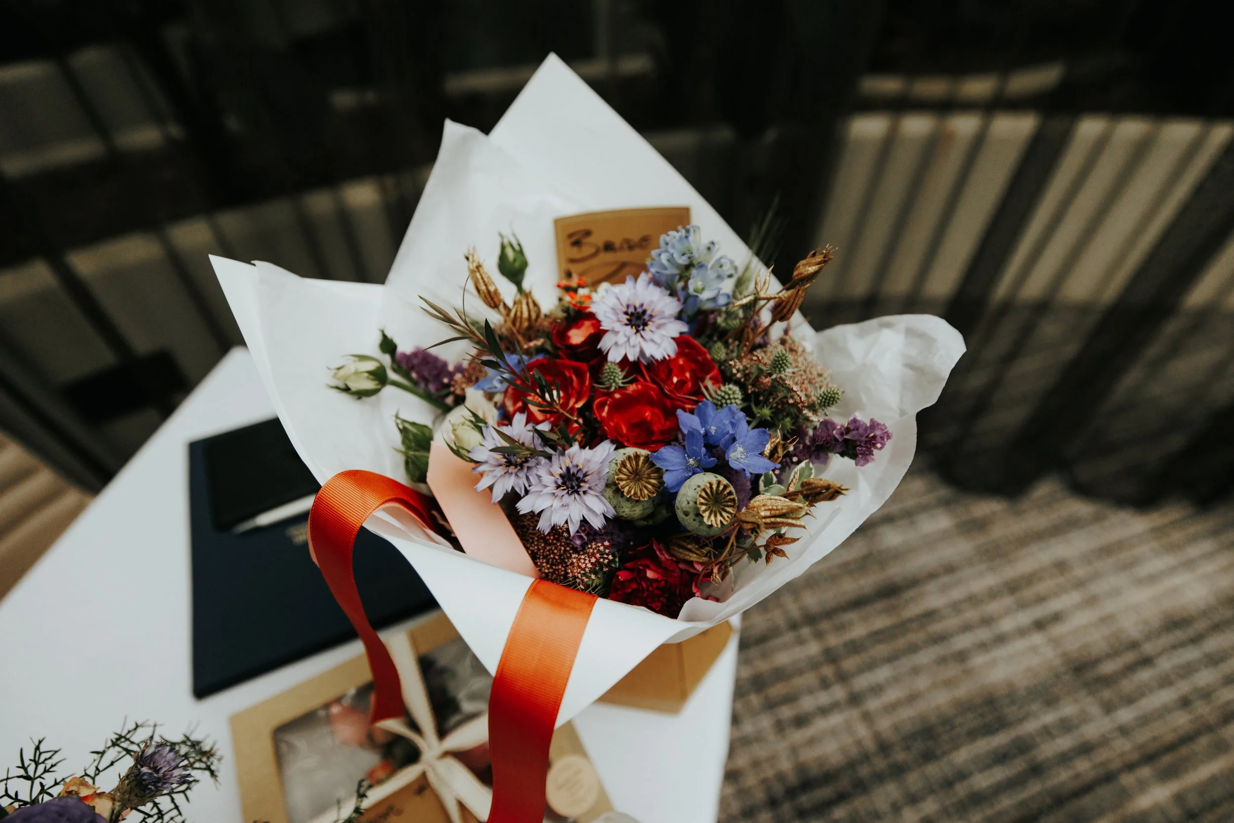 Bouquet of colorful flowers with red, blue, and purple blooms wrapped in white paper with an orange ribbon on a table.