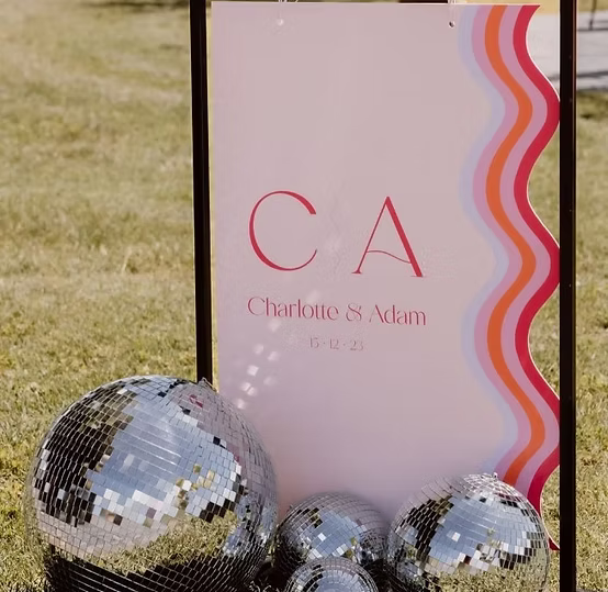 Wedding sign with initials "CA" and date, surrounded by disco balls, placed on grass.