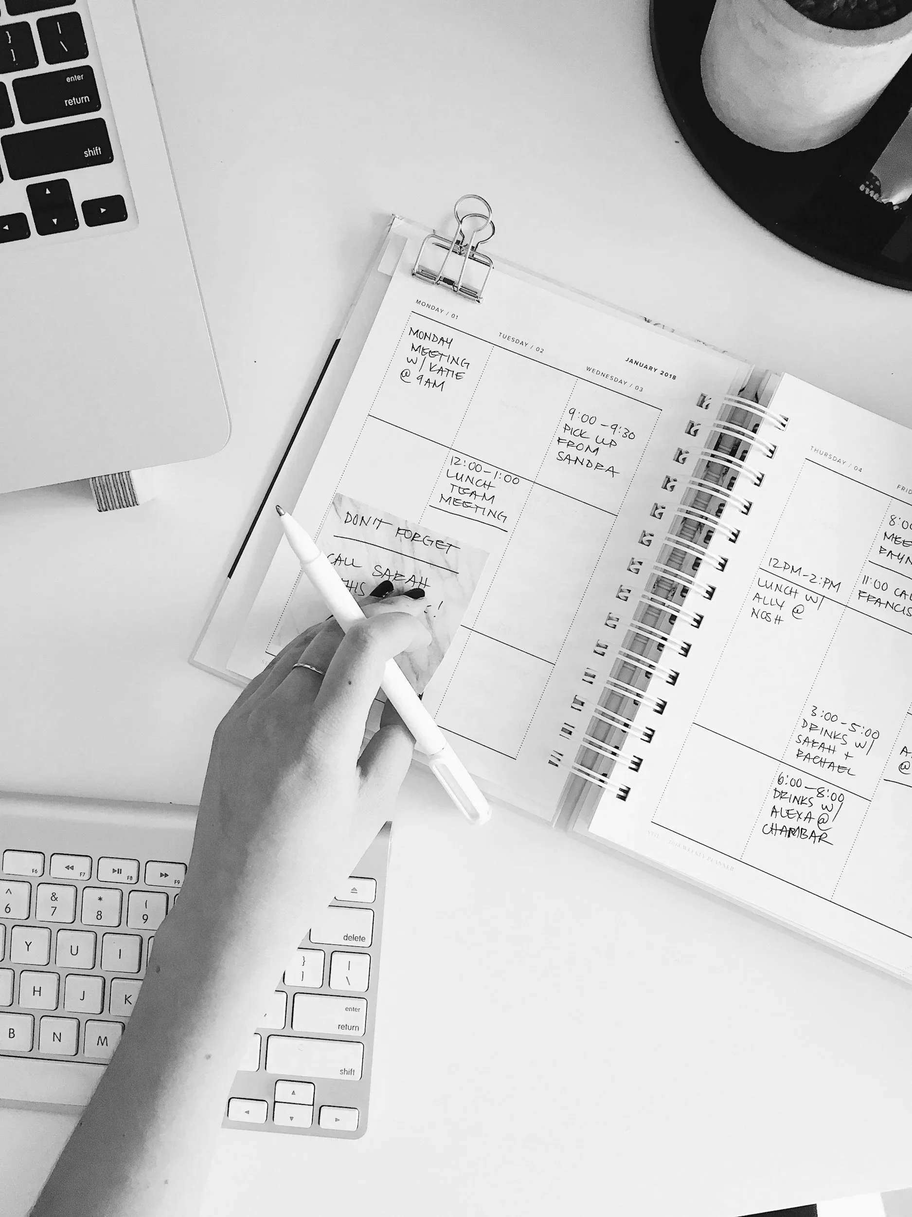 An open planner on a desk with handwritten appointments, a hand holding a white pen, a keyboard, a laptop, and a plant in a pot.