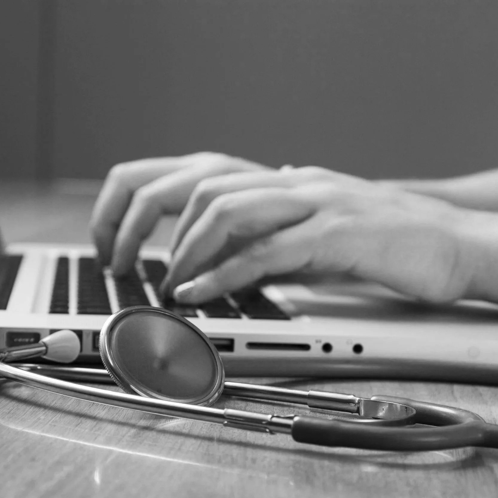 Black and white photograph of a medical stethoscope resting on a wooden surface, with a laptop keyboard and a person's hand in the background.
