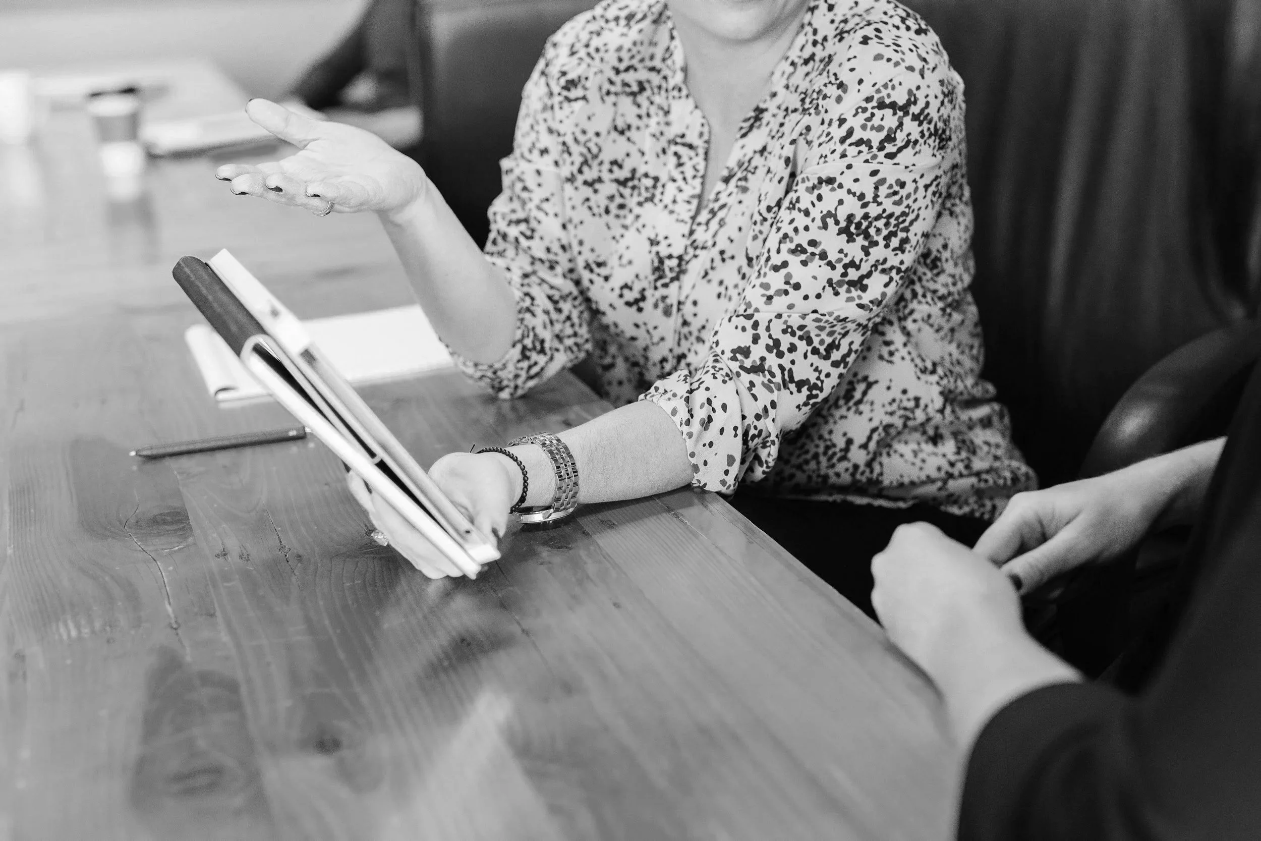 A woman, a midwife, in a patterned blouse, is sitting at a table, gesturing with her left hand while speaking to someone- her patient. There are notebooks and a pen on the table, and other people are partially visible.
