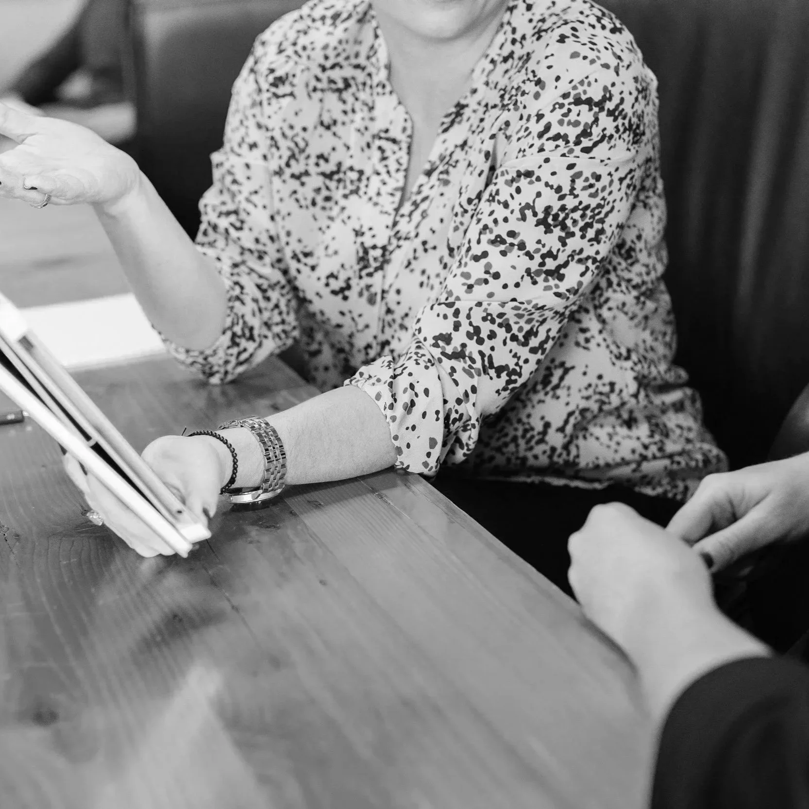 A woman with light skin wearing a patterned blouse sitting at a wooden table, with her arm extended and hand clasped by another person with light skin, whose hand is visible in the foreground.