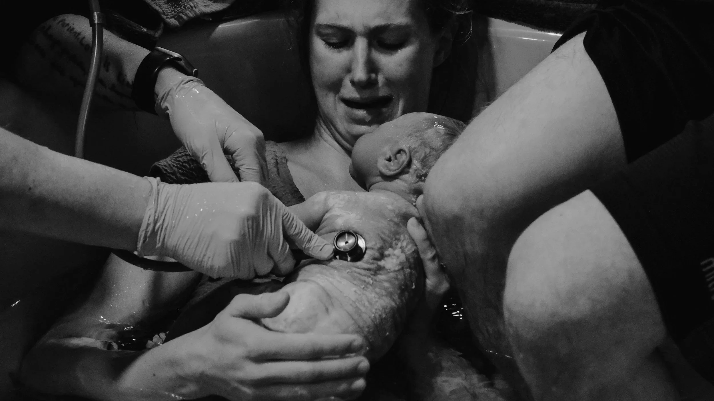 Black and white photo of a woman giving birth at home, surrounded by a midwife and birth assistant, maybe even a doula,  assisting with the delivery.